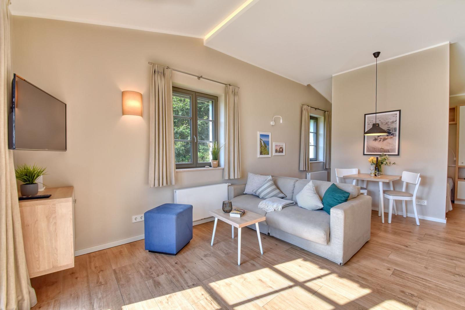 Wohn- und Essbereich mit Sofa, Tisch und Stühlen. Fenster mit Vorhängen und Holzboden.
