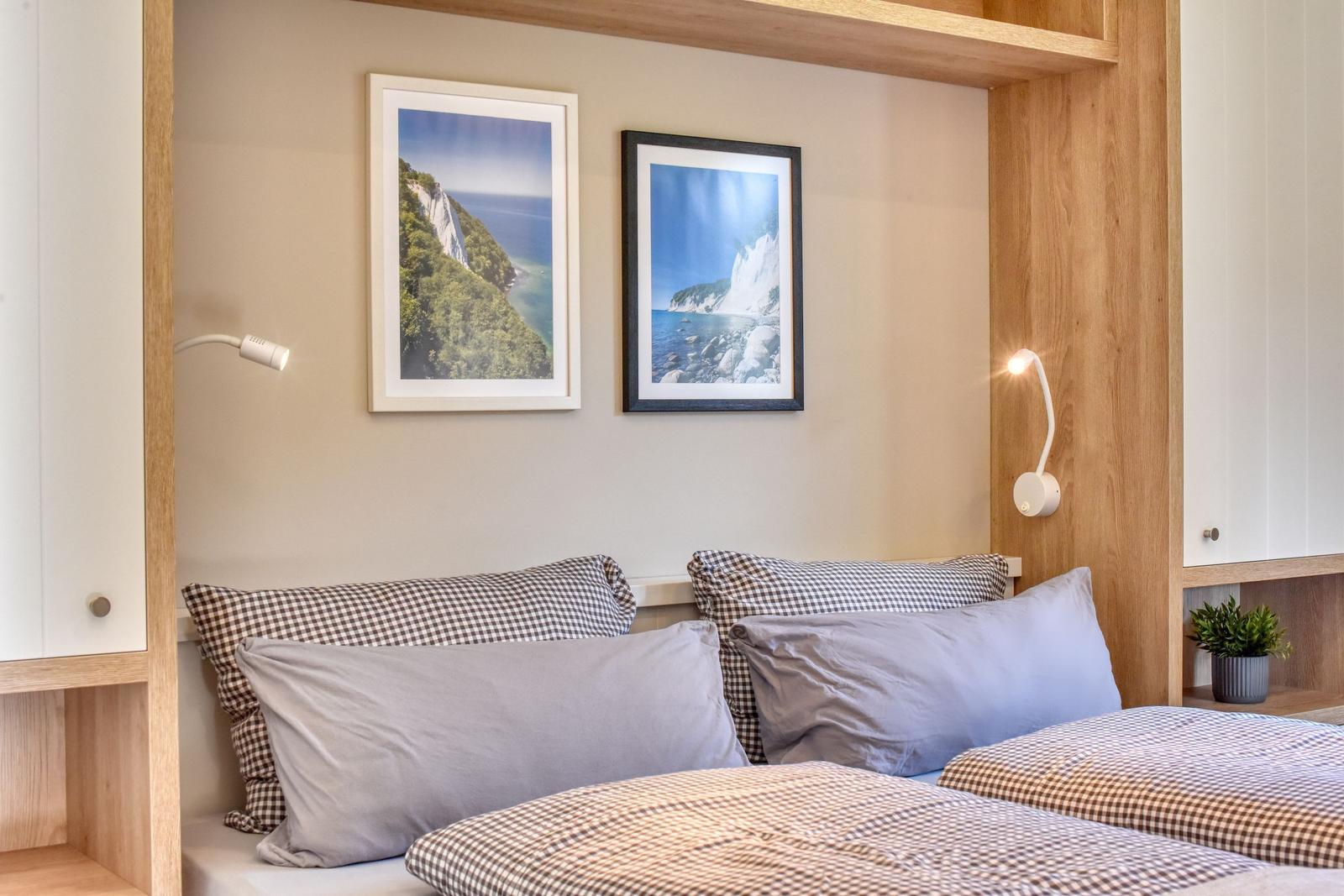 Schlafzimmer mit Bett, Kissen und Wandbildern. Beleuchtung und Pflanze sichtbar.