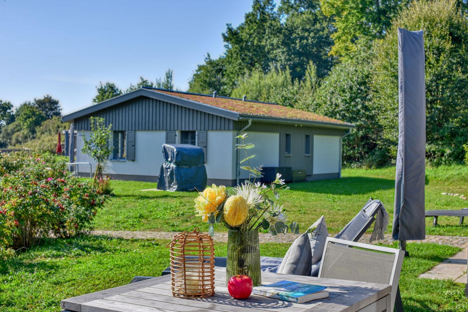 Terrasse mit Tisch, Stühlen und Blumen. Im Hintergrund ein Haus mit Garten.