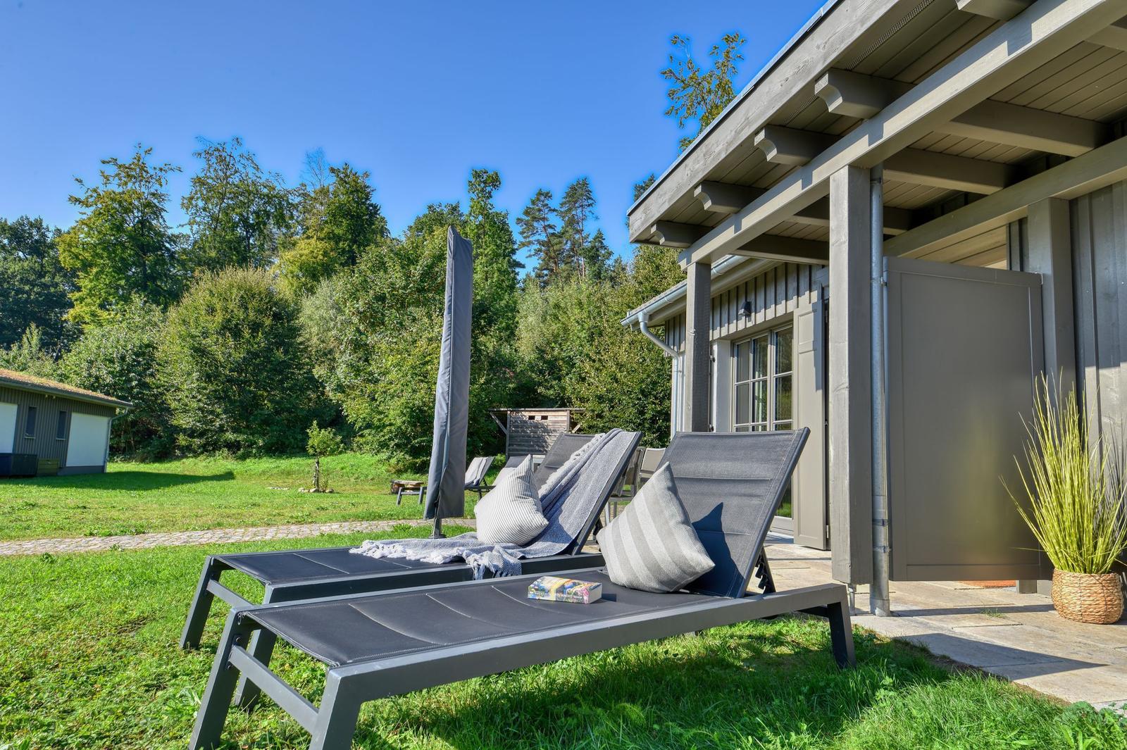 Zwei Liegestühle mit Kissen stehen auf einer Terrasse vor einem Haus. Hintergrund: grüner Garten und Wald.