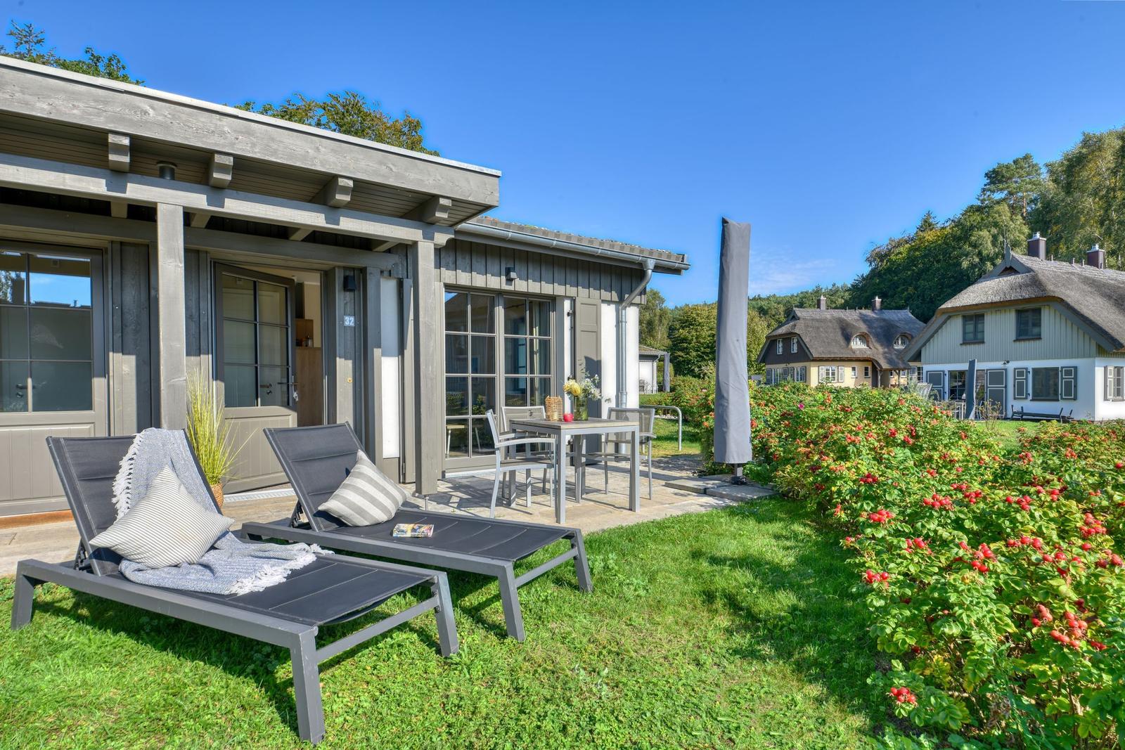 Garten mit Liegen, Tisch und Stühlen vor einem Holzhaus. Rosenhecke und weitere Häuser im Hintergrund.