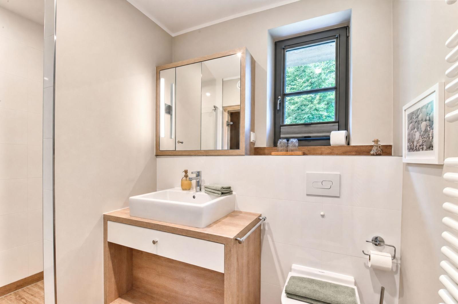 Badezimmer mit Waschtisch, Spiegel und Fenster mit Blick auf Grün.