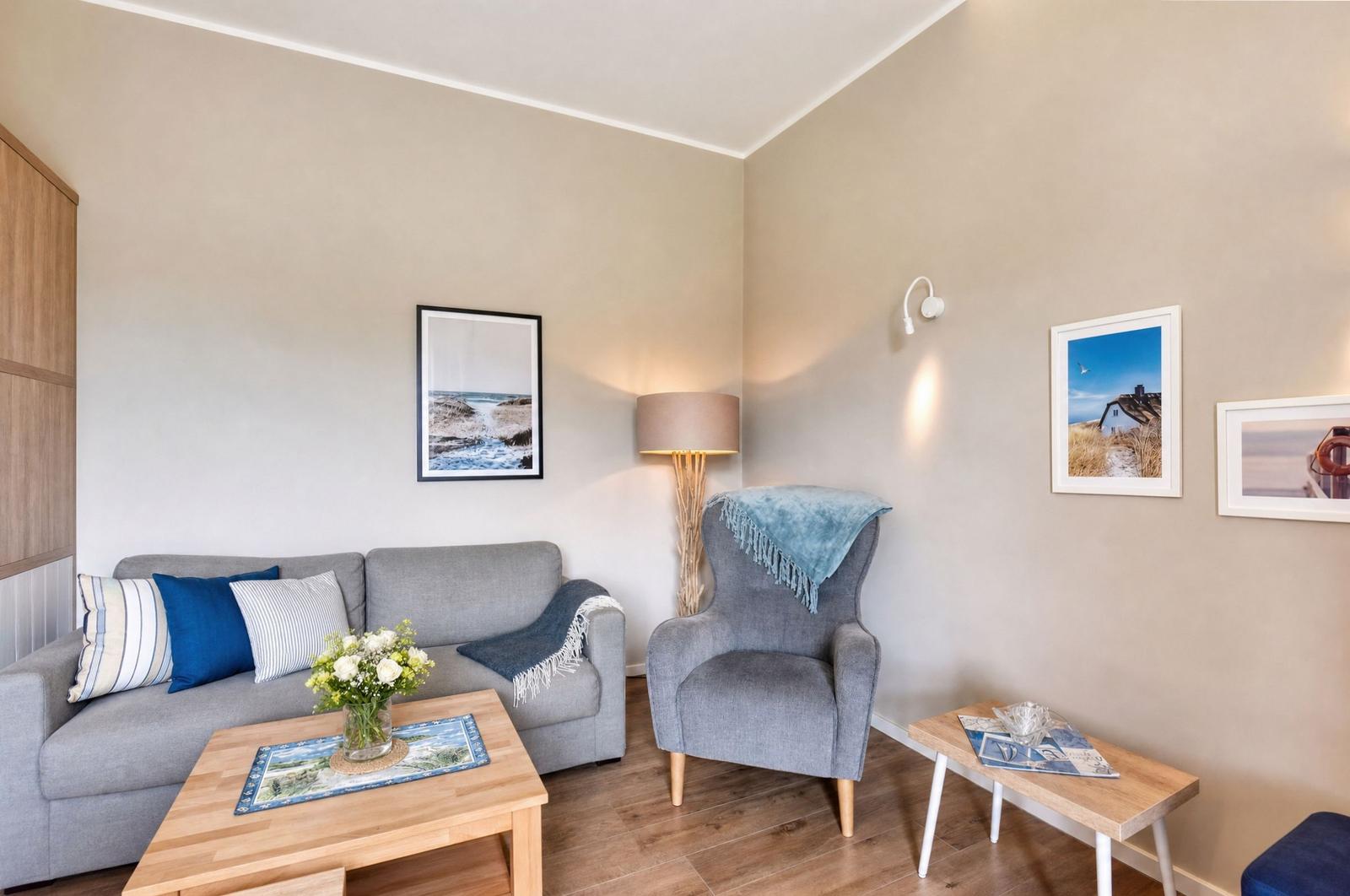 Ein Wohnzimmer mit grauem Sofa, Sessel und Holztisch. An den Wänden hängen Bilder.