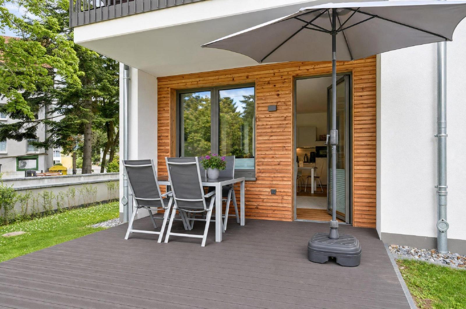 Terrasse mit Tisch, Stühlen und Sonnenschirm. Durchgang in die Küche. Holzwand und grüner Garten.