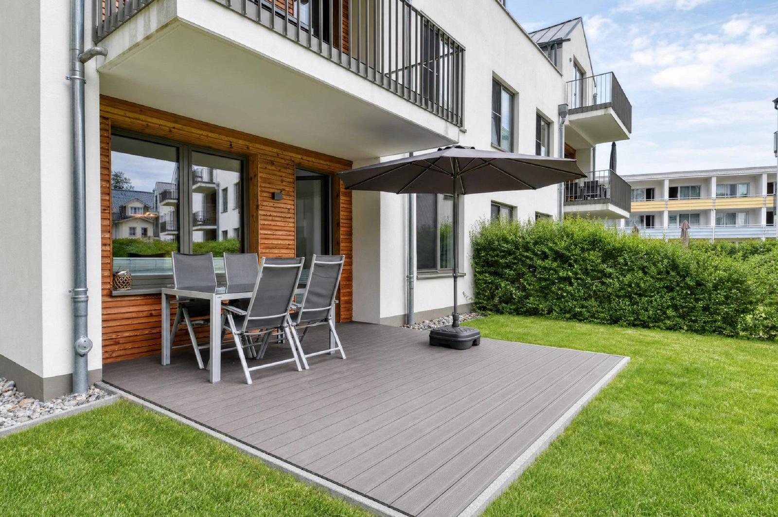 Terrasse mit Tisch, vier Stühlen und Sonnenschirm. Grünfläche und Hecke im Hintergrund.
