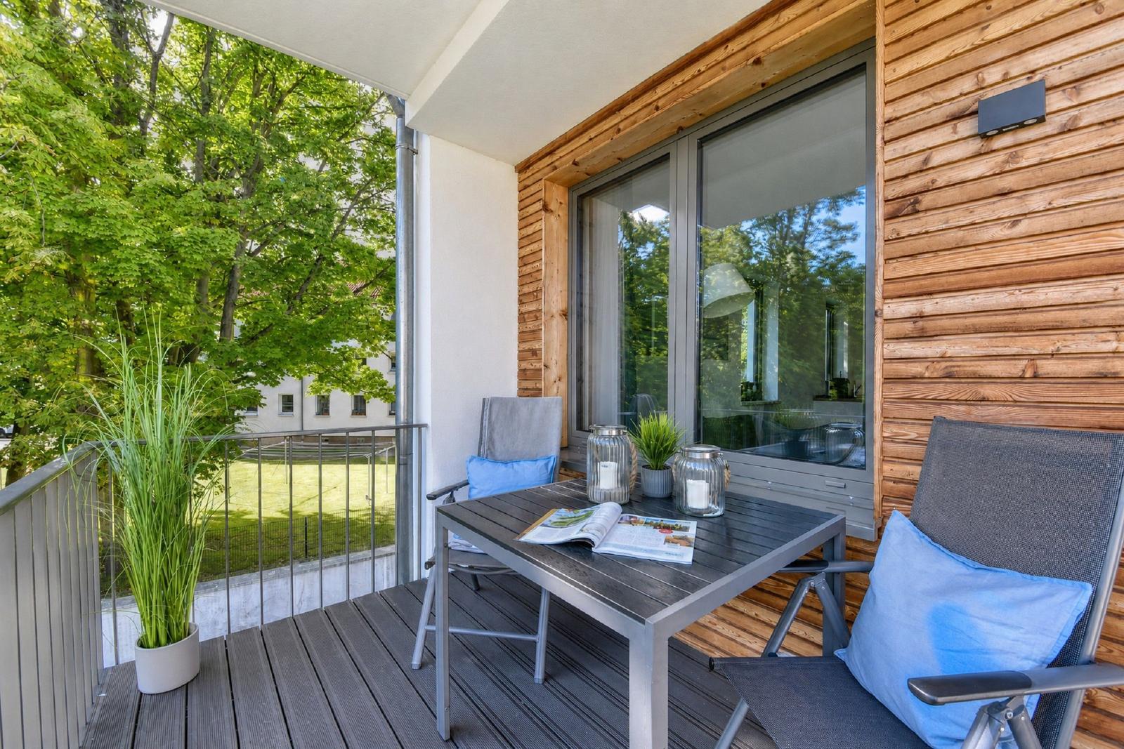 Terrasse mit Tisch, zwei Stühlen und Pflanzen. Holzwand und Glasfenster sichtbar.