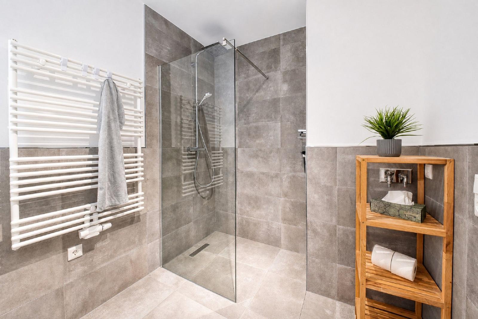Dusche mit Glaswand, Heizkörper und Holzregal mit Handtüchern.