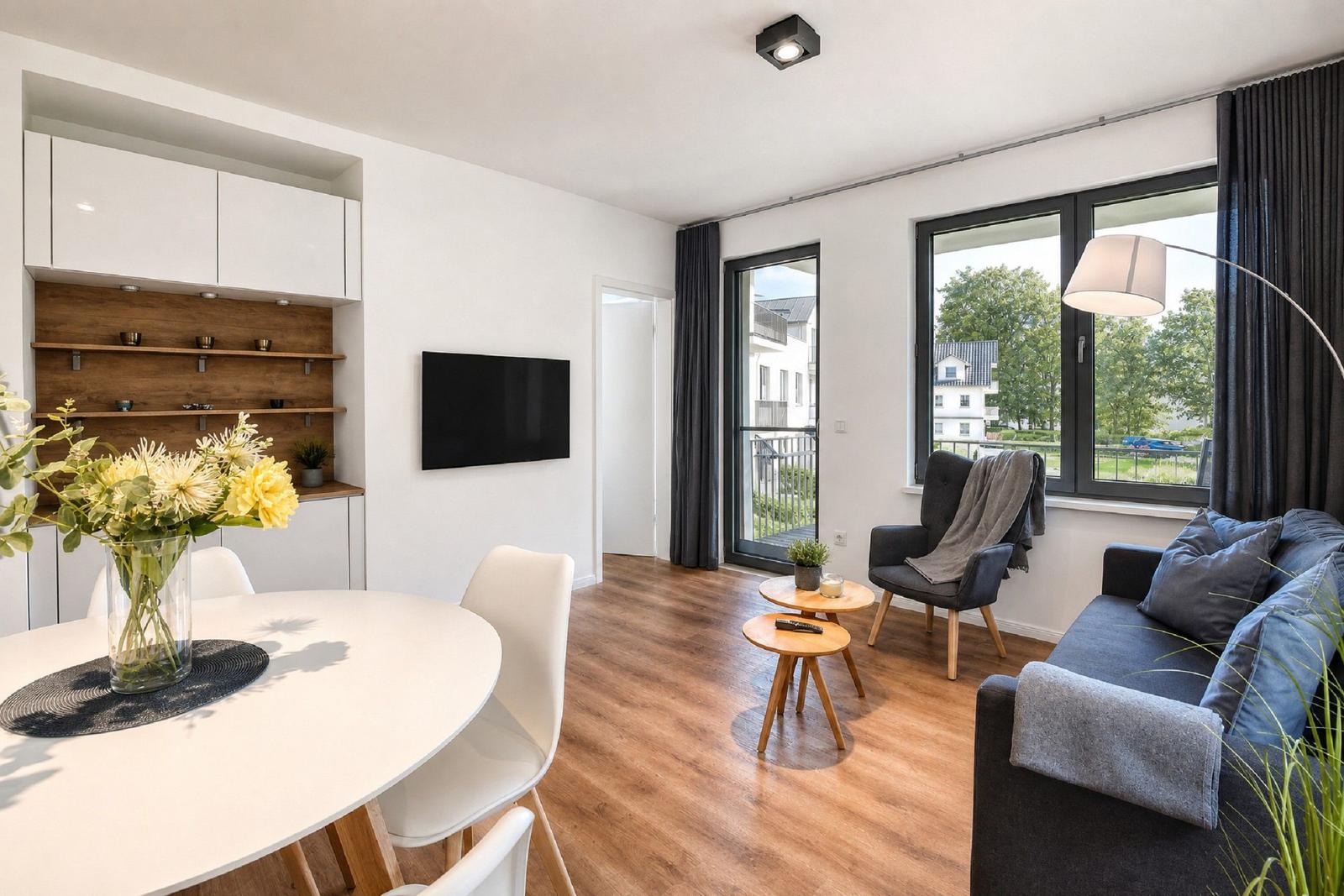 Wohn- und Essbereich mit Sofa, Stuhl, Tisch und Fernseher. Holzboden und große Fenster.
