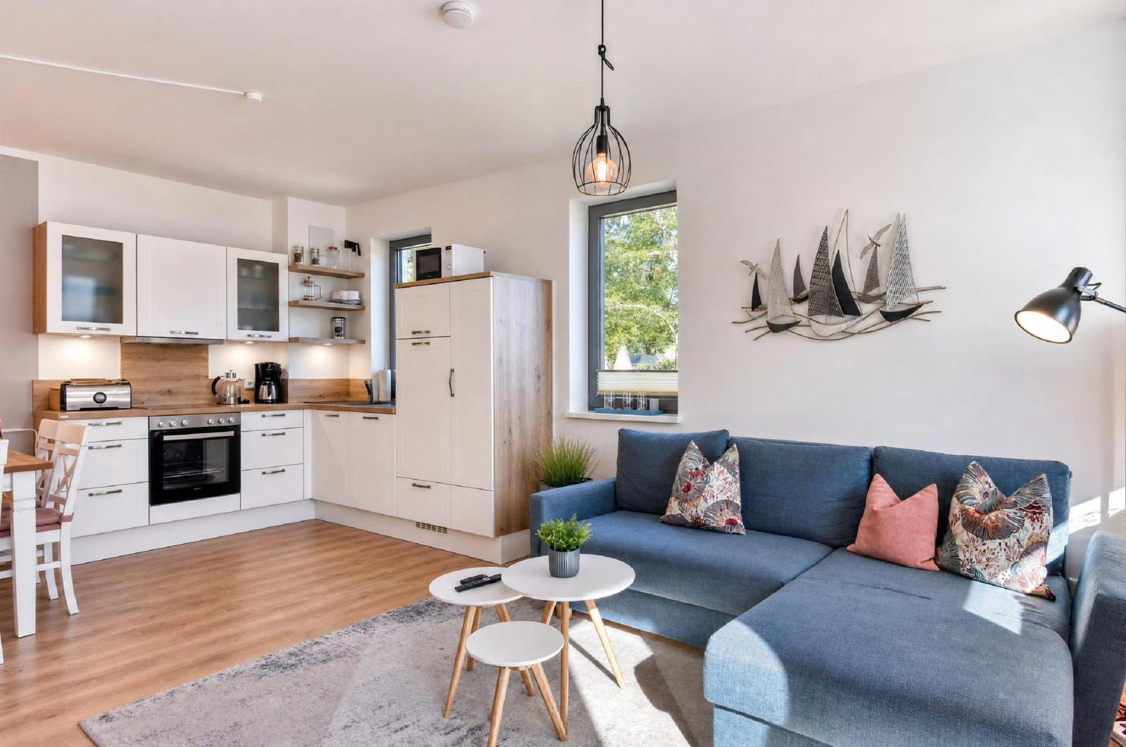 Wohnbereich mit Sofa, Tisch und Küchenzeile. Wanddekoration und Fenster sichtbar.