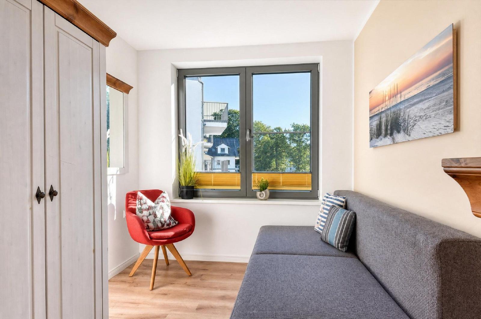 Ein Wohnzimmer mit grauem Sofa, roter Sitzgelegenheit und Fensterblick auf Bäume.