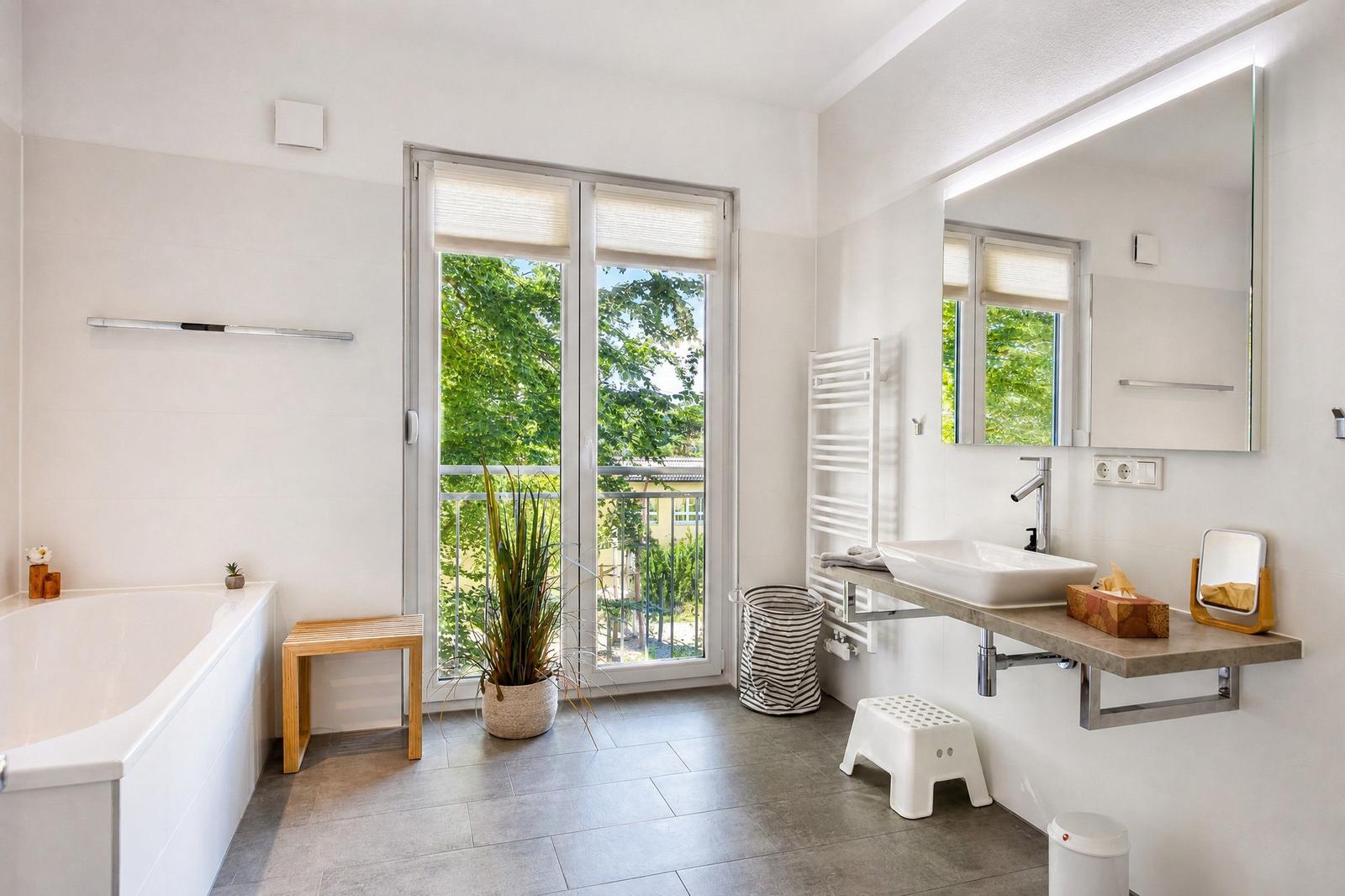 Badezimmer mit Badewanne, Waschbecken und Balkon mit Blick auf Grünflächen.