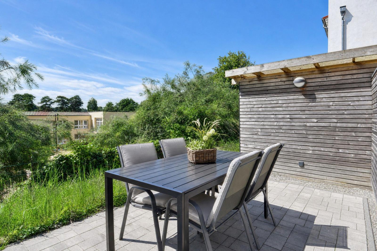 Terrasse mit Tisch und Stühlen, Holzwand und Grünflächen im Hintergrund.