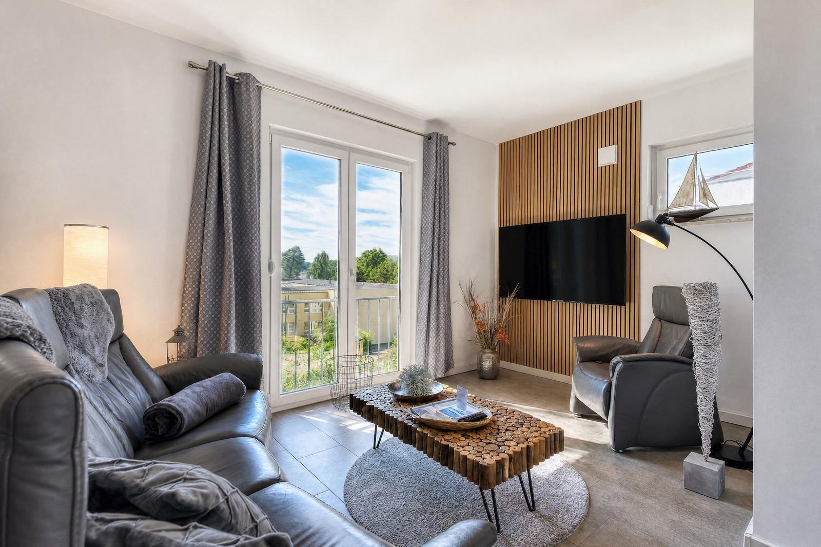 Wohnzimmer mit Ledersofa, Holztisch und TV an Holzwand. Balkon mit Blick auf Grünflächen.