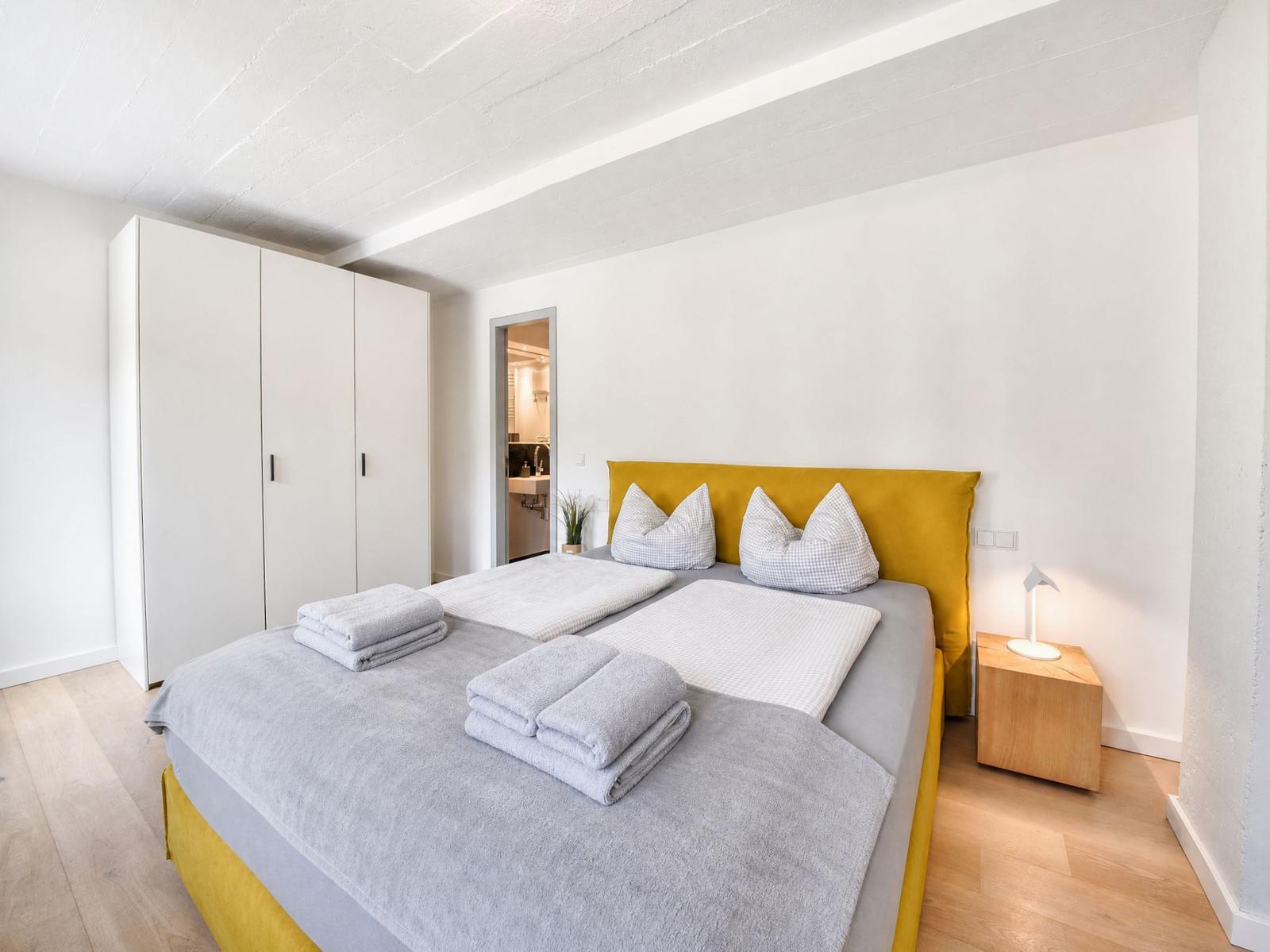 Schlafzimmer mit gelbem Bett, weißen Wänden und Holzboden. Neben dem Bett steht ein Nachttisch.
