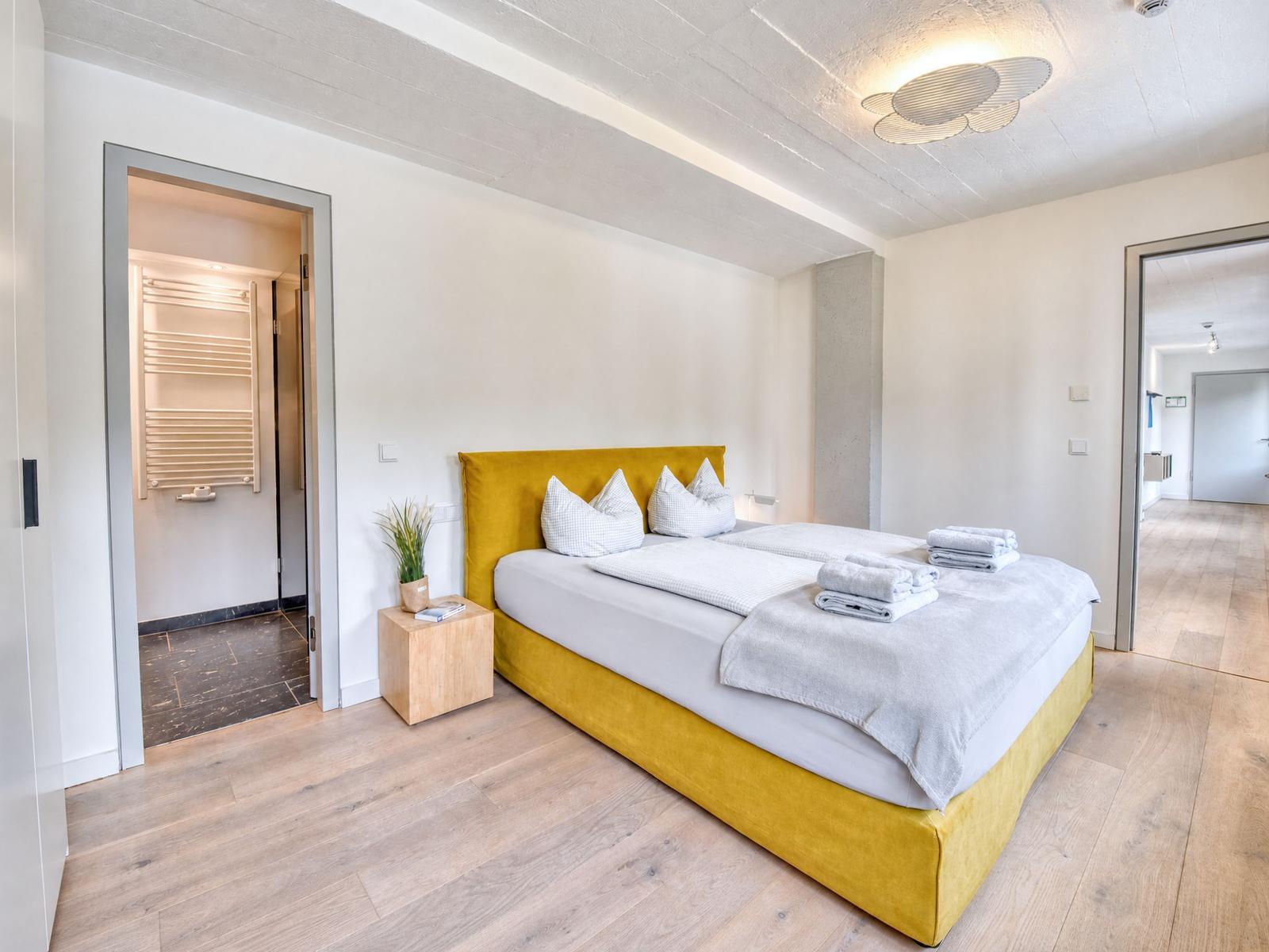 Schlafzimmer mit gelbem Bett, Holzboden und Blick in Bad und Flur.