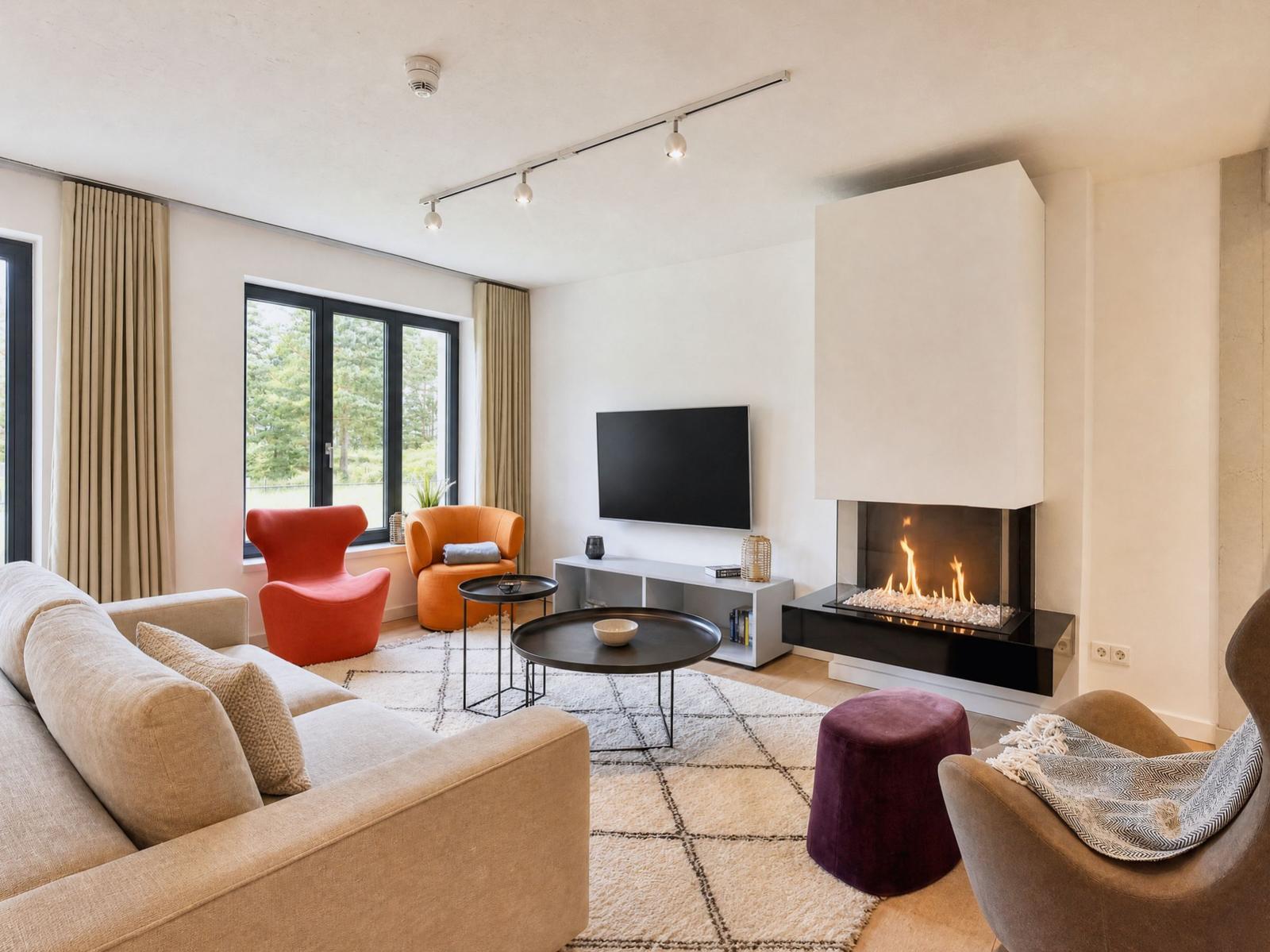 Wohnzimmer mit Sofa, Sesseln, Kamin und Fernseher. Fenster mit Blick auf Grün.