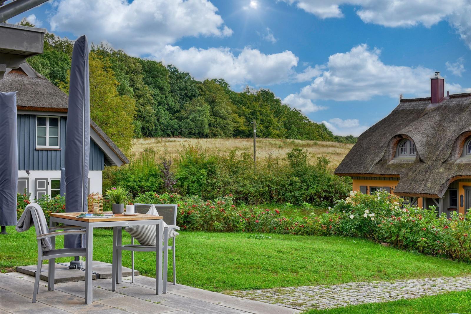 Terrasse mit Tisch und Stühlen, Blick auf grünen Garten und strohgedeckte Häuser.