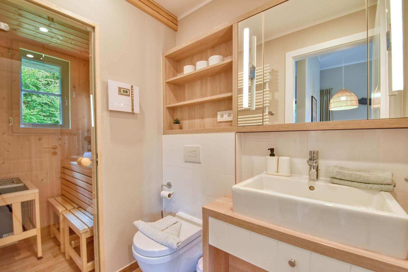 Badezimmer mit Dusche, Waschbecken und Toilette. Holzregale und Spiegel sind vorhanden.