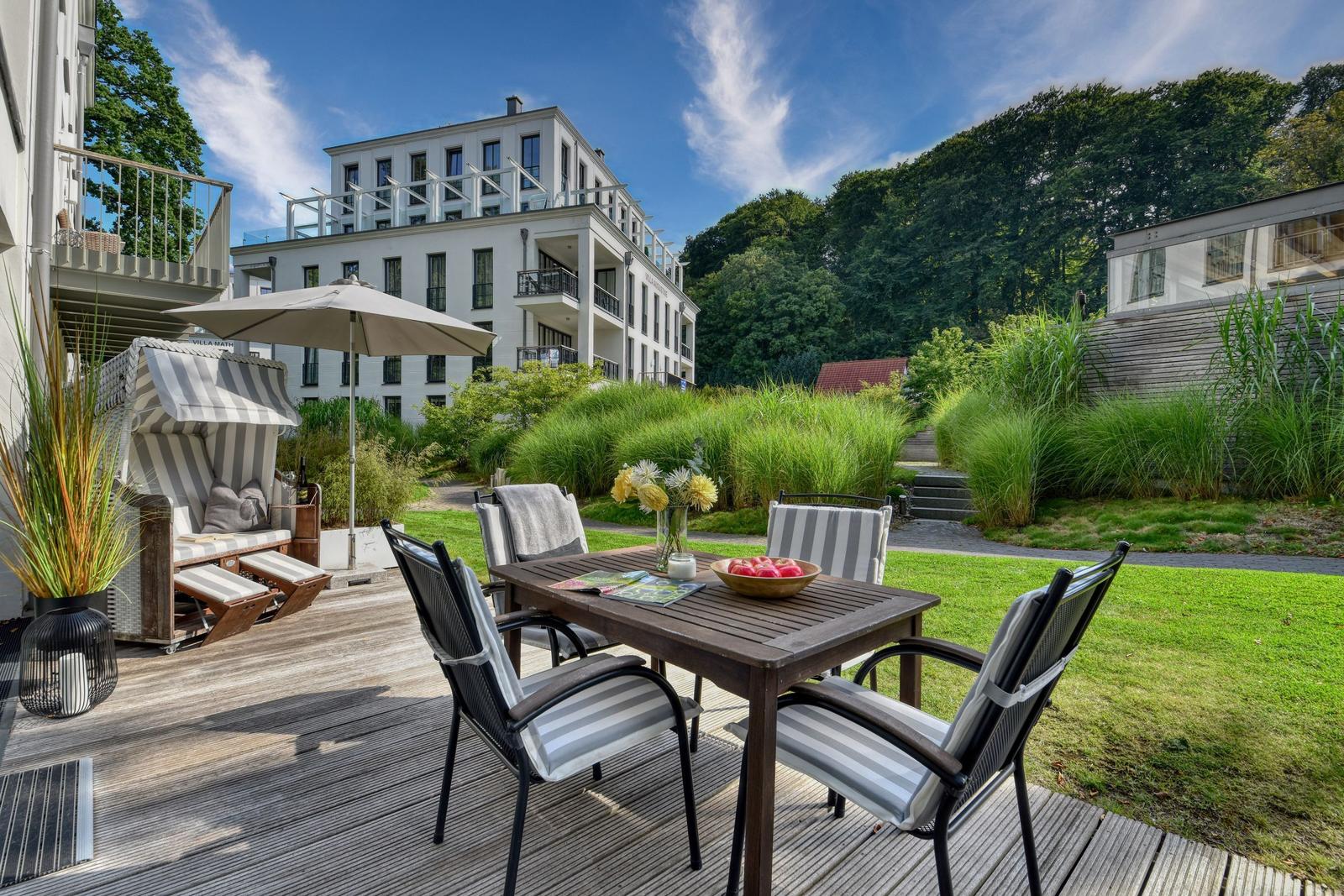 Terrasse mit Tisch, Stühlen und Sonnenschirm. Hintergrund: modernes Gebäude und Grünflächen.