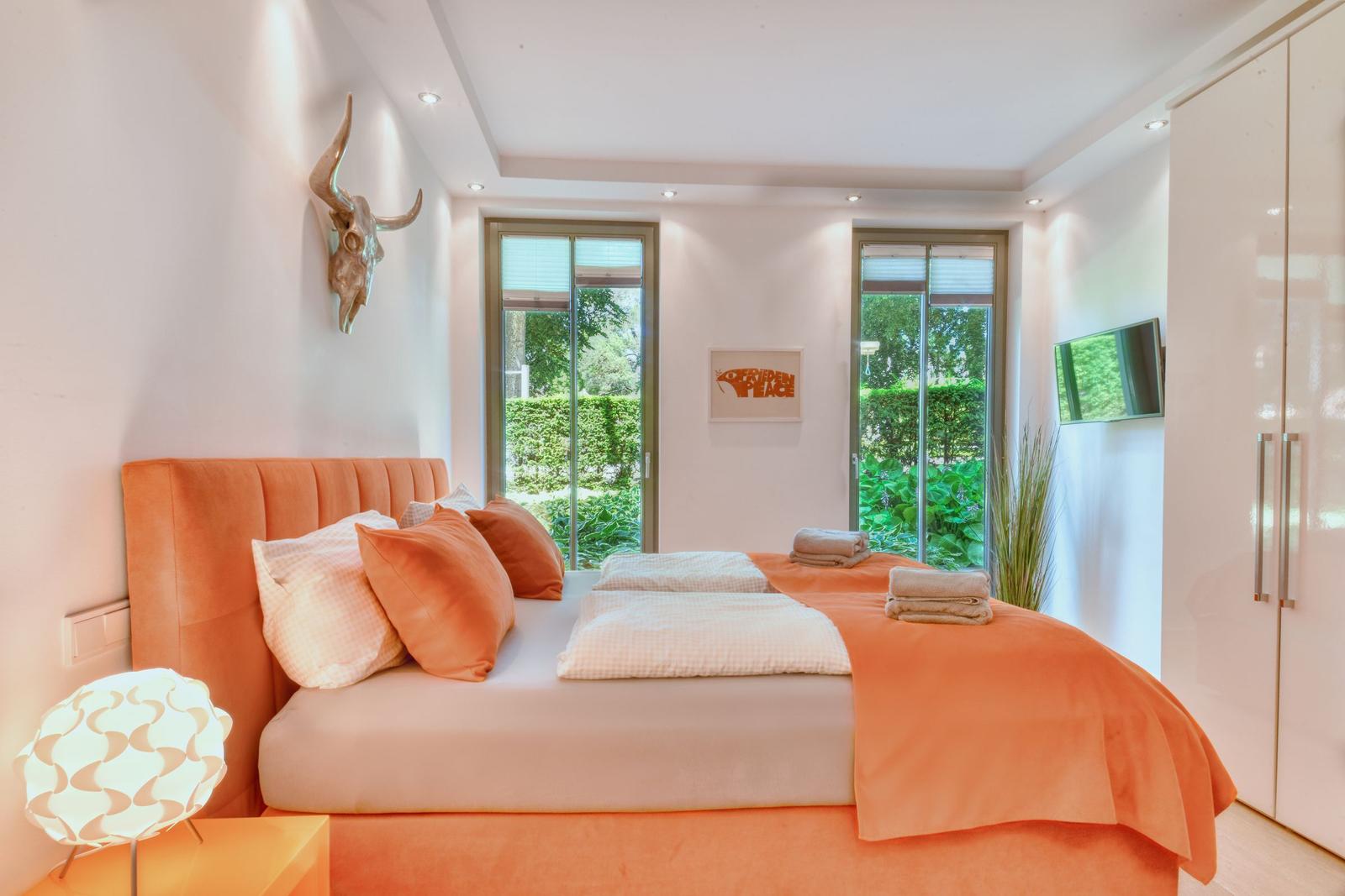 Schlafzimmer mit orangeem Bett, Fenstern und Wandgarderobe