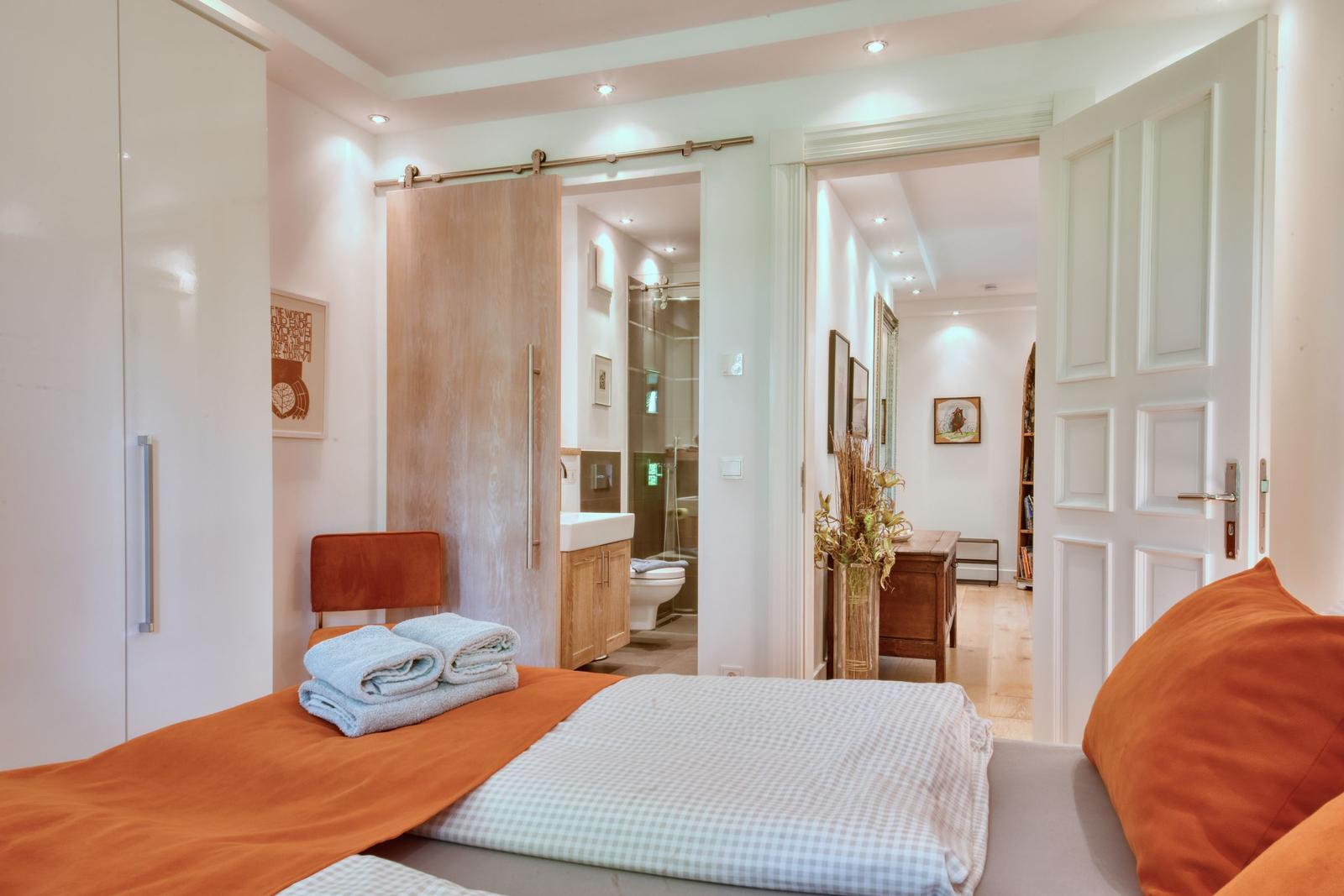Schlafzimmer mit Bett, orange Bettwäsche und Blick in Bad und Flur.