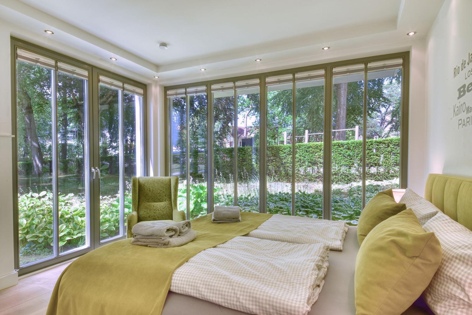 Schlafzimmer mit großem Fensterblick auf Garten, Bett mit grünen Kissen und Decke.