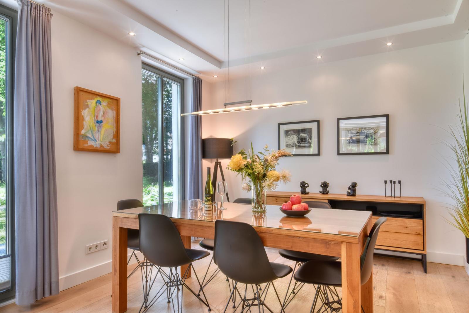 Esszimmer mit Holztisch, schwarzen Stühlen und Glasplatte. Fenster, Wandbilder und Pflanzen.