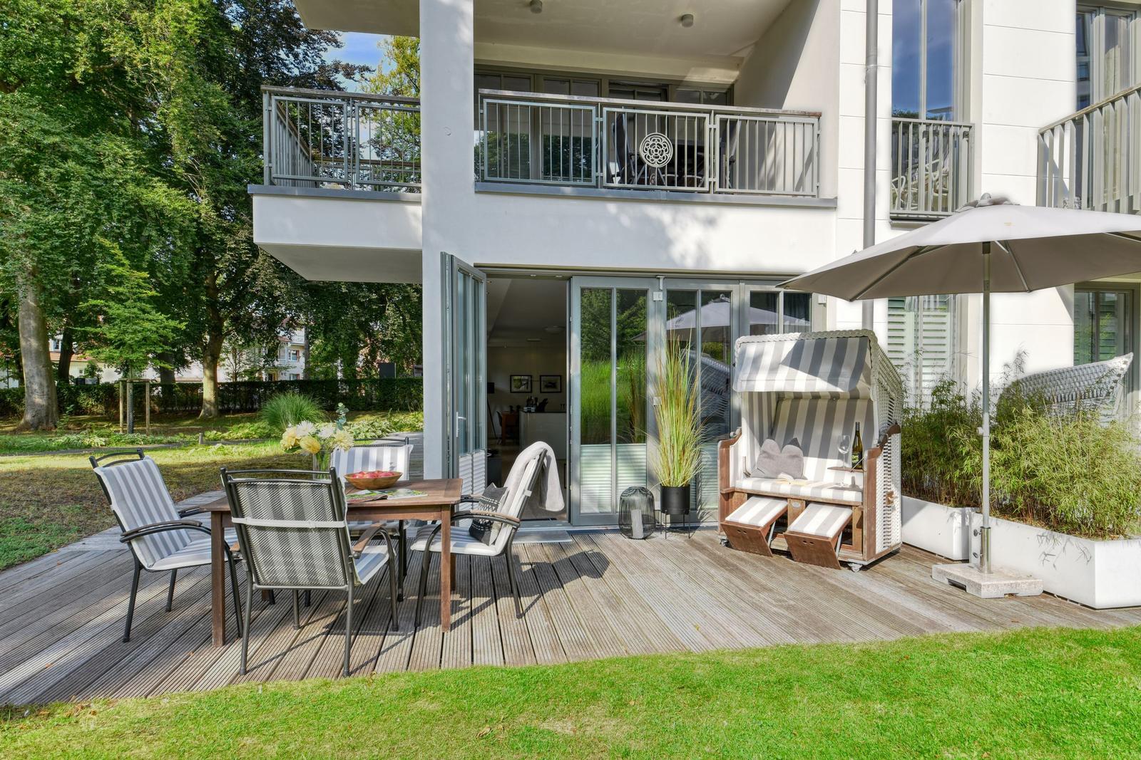 Terrasse mit Tisch, Stühlen und Liege unter Sonnenschirm. Hinterhof mit Grünfläche und Balkon.