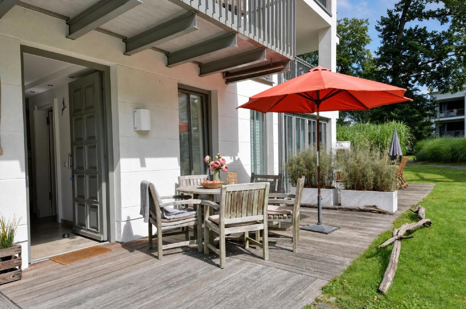 Terrasse mit Tisch, Stühlen und rotem Sonnenschirm. Hinterhof mit Gras und Pflanzen.