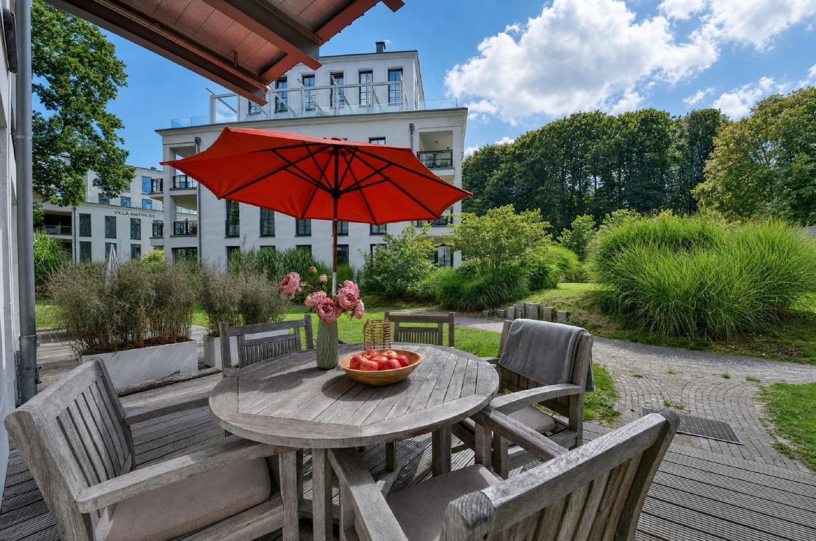 Terrasse mit Holztisch, Stühlen und rotem Sonnenschirm. Im Hintergrund Villa Mathilde und Grünflächen.