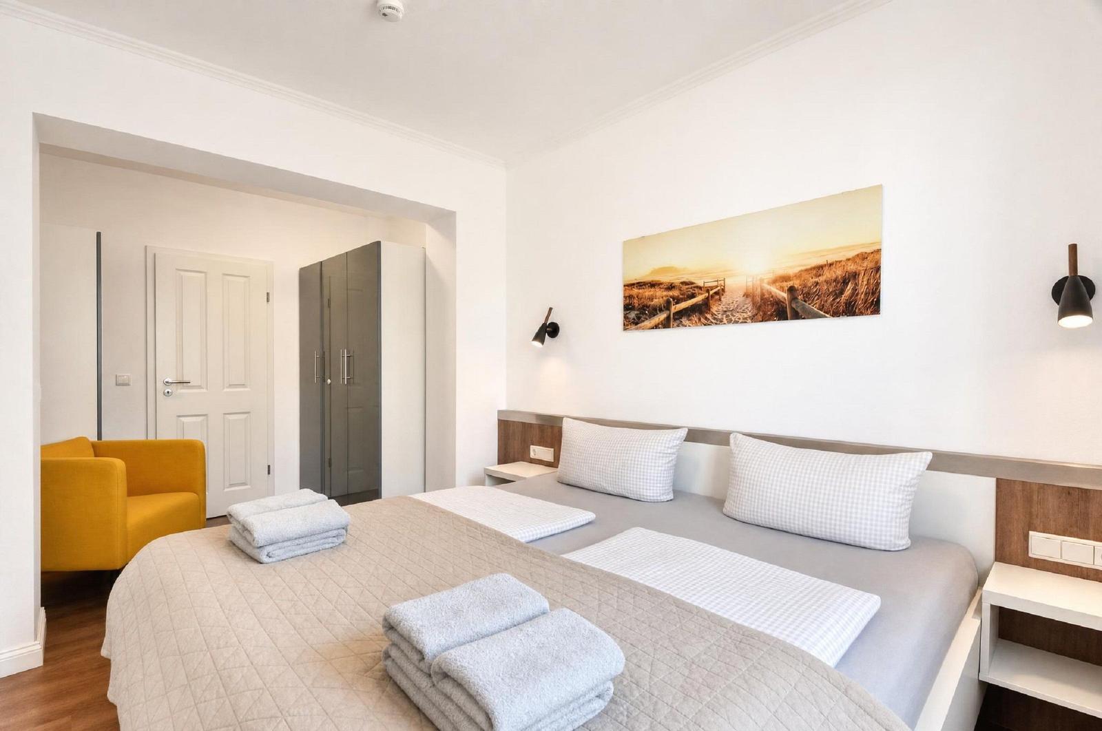 Schlafzimmer mit Doppelbett, Wandbild und Nachttischen. Gelber Sessel und Kleiderschrank sichtbar.