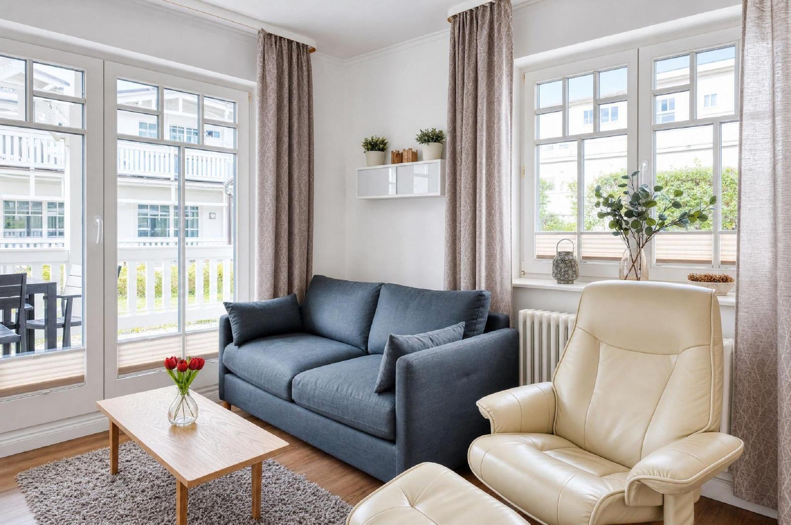 Wohnzimmer mit grauem Sofa, cremefarbenem Sessel und Holztisch. Große Fenster und Balkontür.