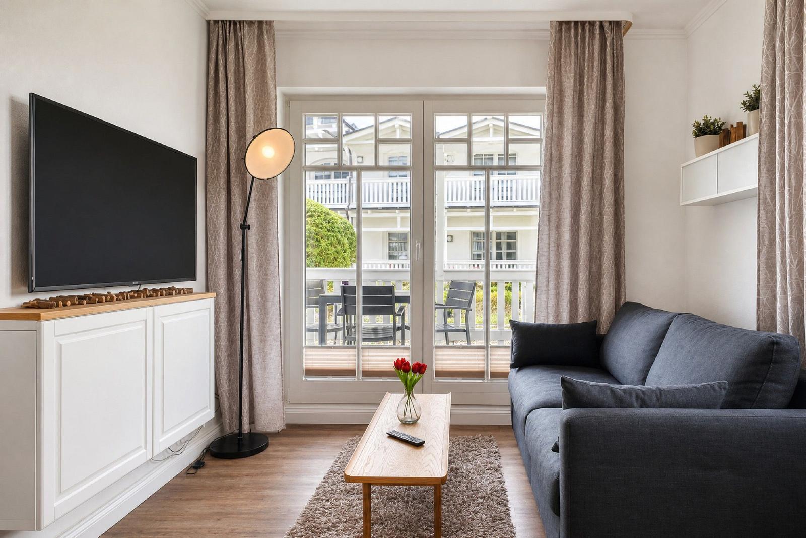 Ein Wohnzimmer mit Sofa, Fernseher und Balkon. Es gibt einen Tisch mit Blumen.