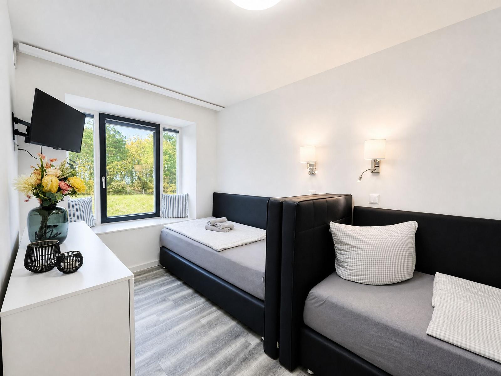 Room with two beds, window with view of green, wall lamp and TV.