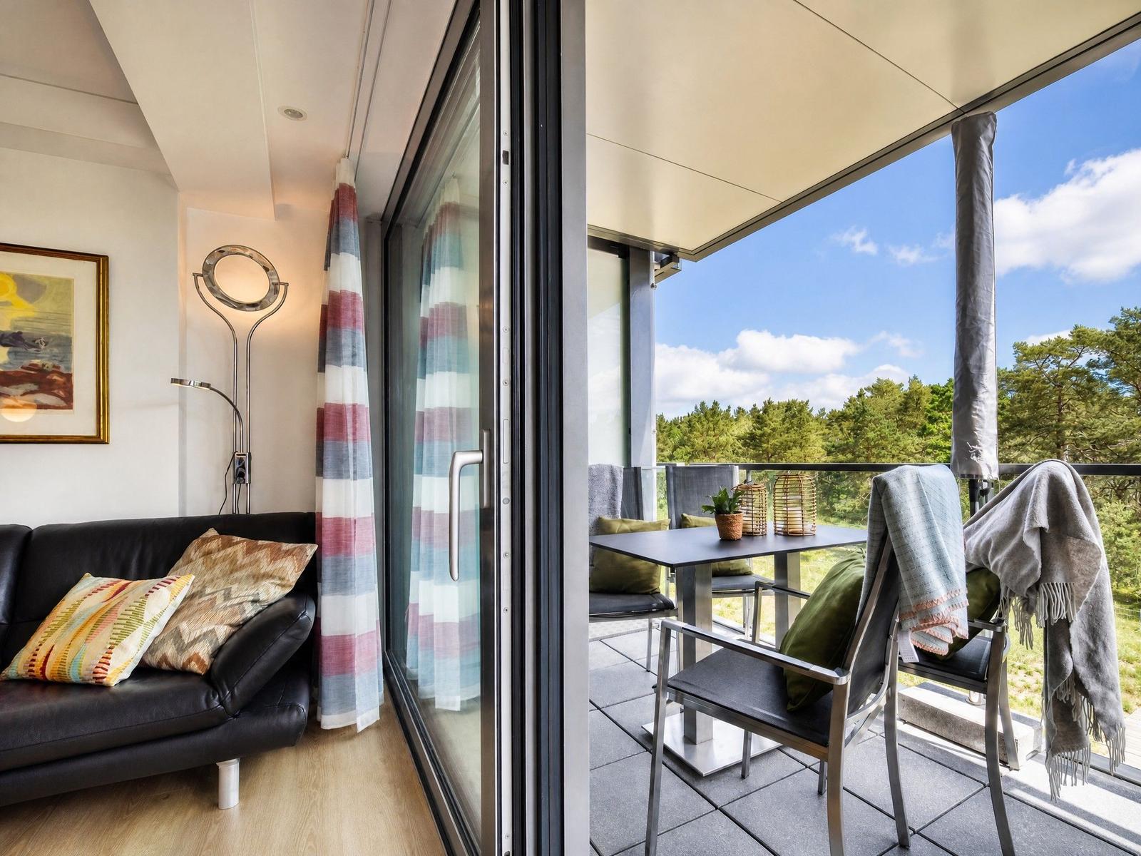 Wohnbereich mit Sofa und Balkon mit Tisch, Stühlen und Blick auf Wald.
