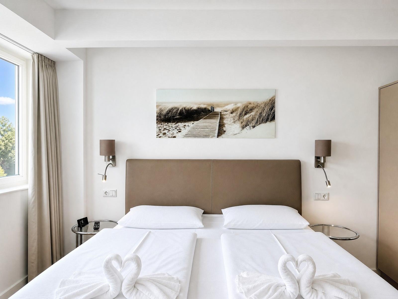 Ein Bett mit weißen Laken und zwei Schwänen aus Handtüchern. An der Wand hängt ein Bild von einer Strandpromenade.
