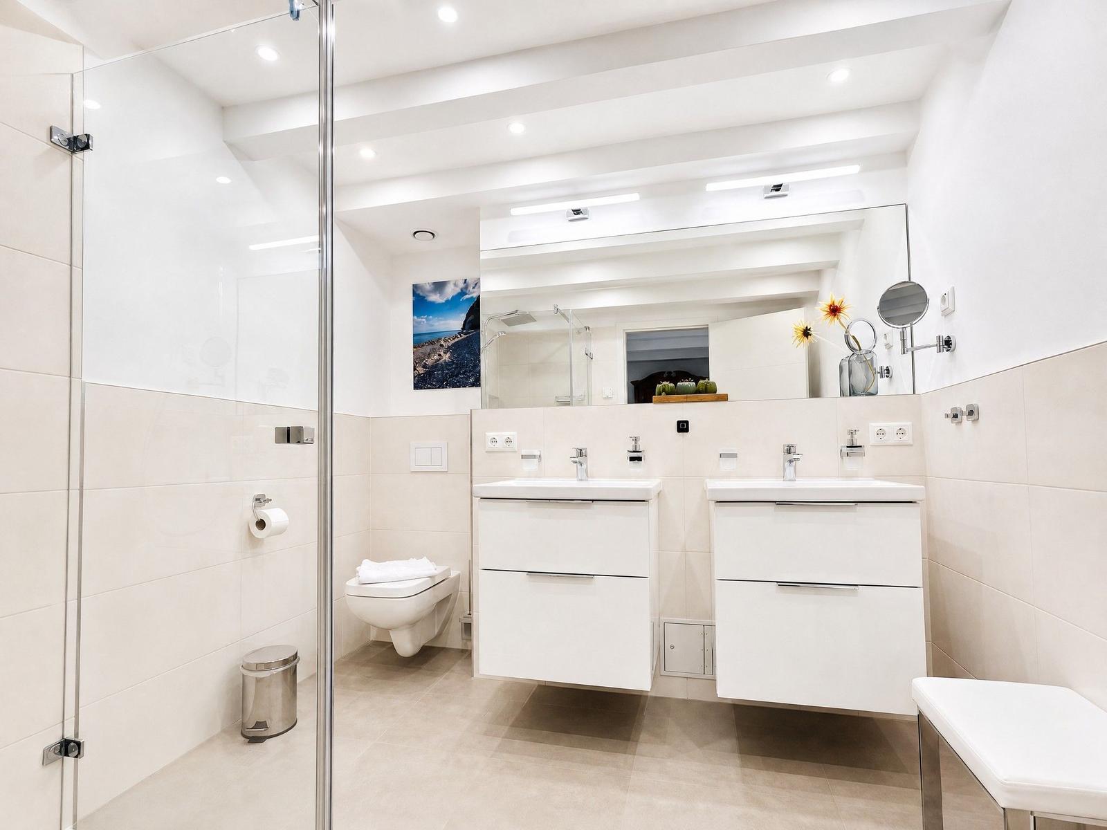 Moderne Dusche mit Glaswand, zwei Waschbecken und Spiegel. Toilette und Sitzbank sind sichtbar.