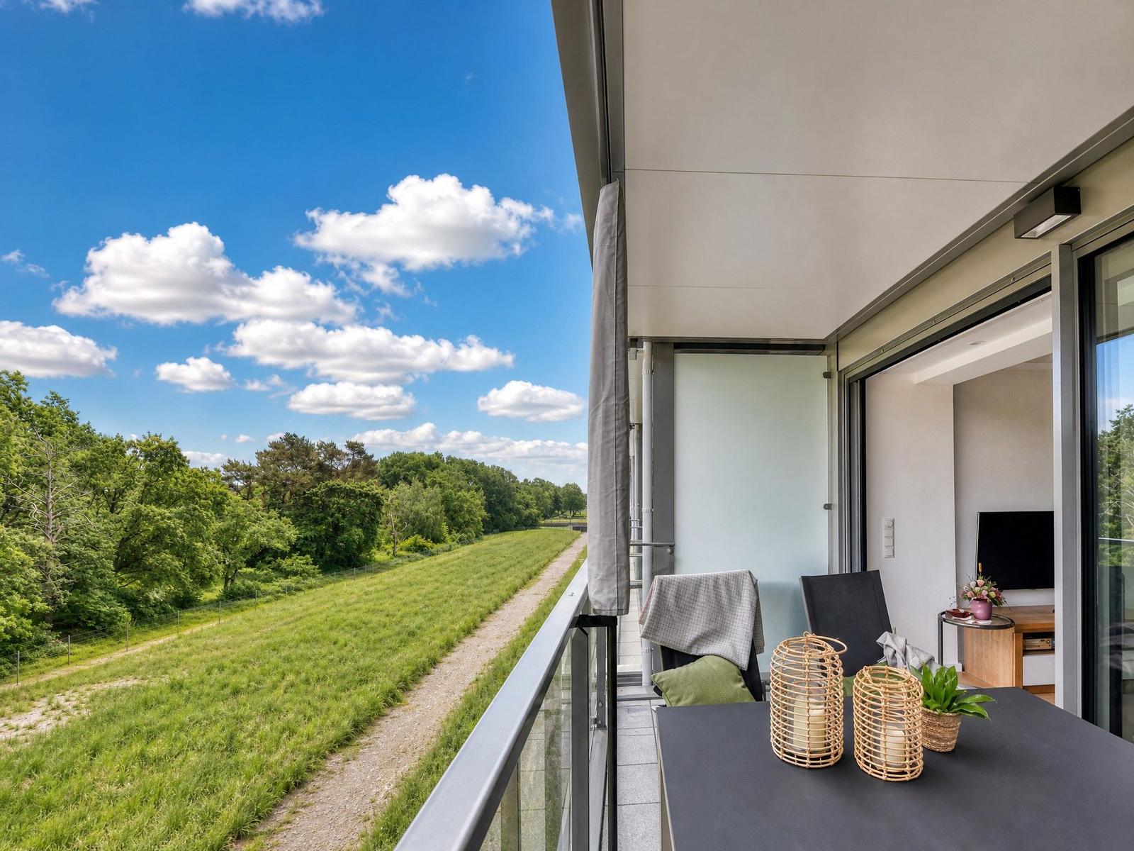 Terrasse mit Blick auf grüne Wiesen und Wald unter blauem Himmel mit weißen Wolken.
