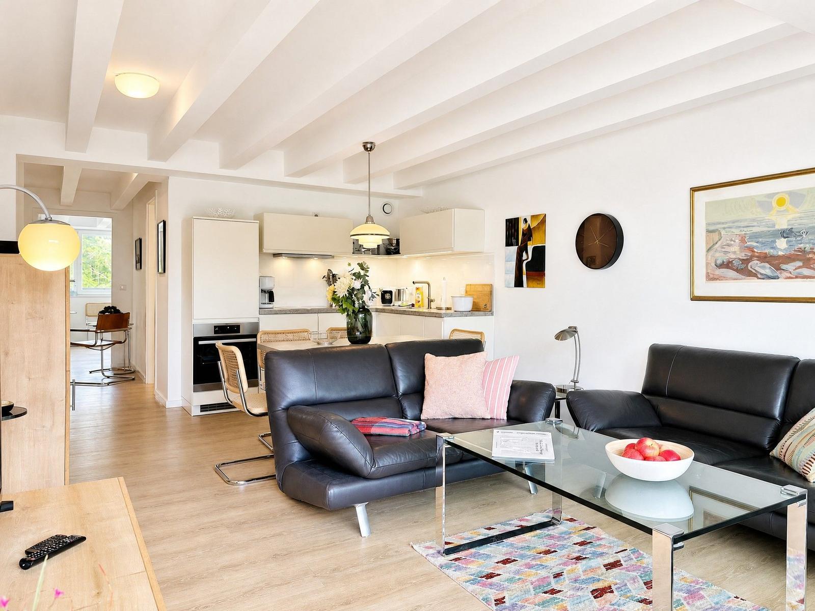 Wohn- und Küchenbereich mit schwarzen Sofas, Glas-Tisch und Holzboden.