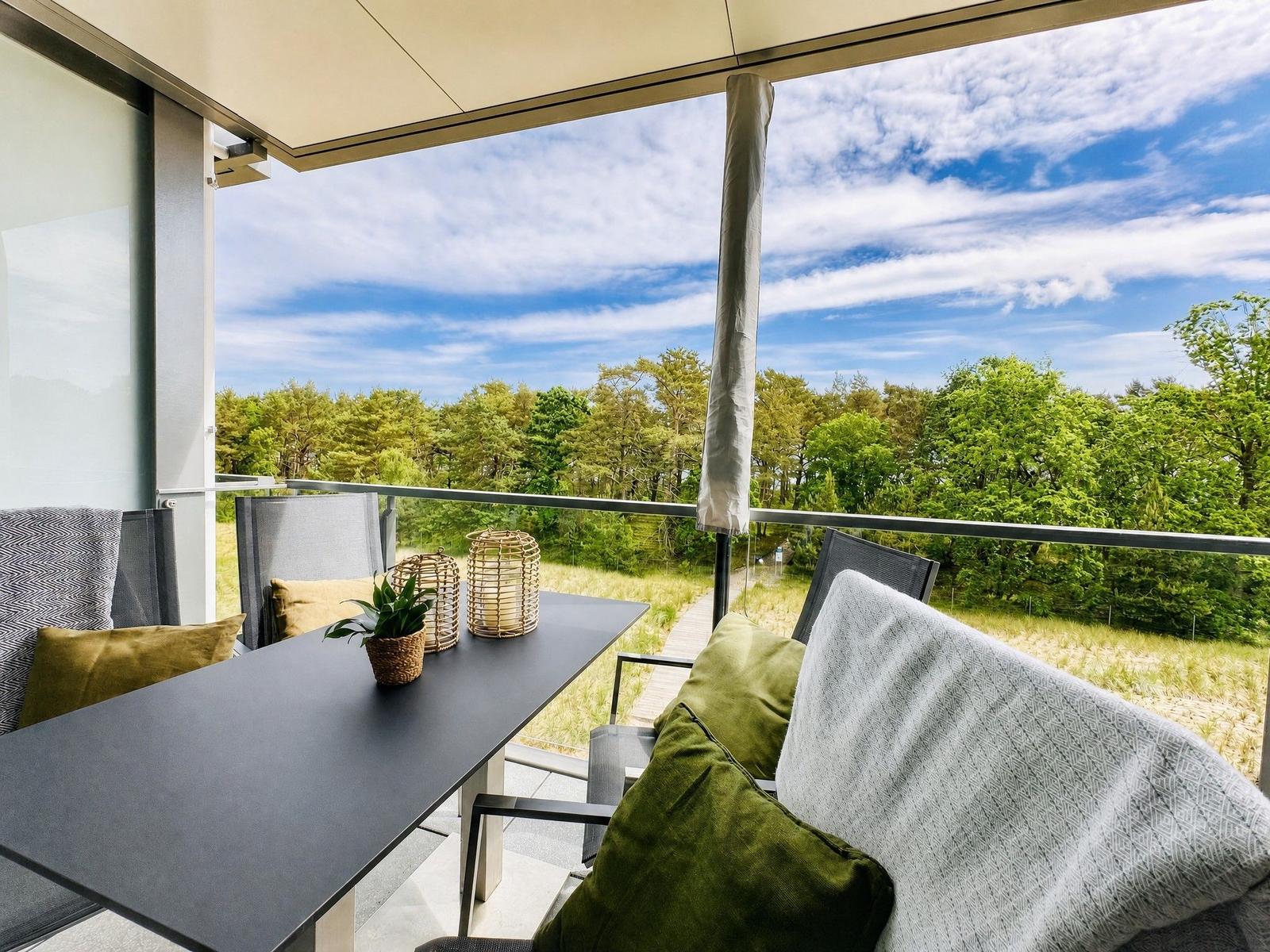 Terrasse mit Tisch, Stühlen und Blick auf Wald. Himmel blau mit weißen Wolken.