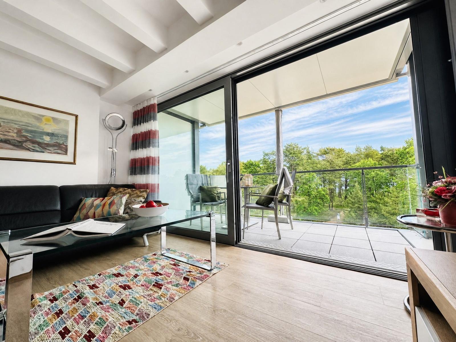 Wohnzimmer mit Balkon und Blick auf Wald. Sofa, Tisch und Teppich sichtbar.