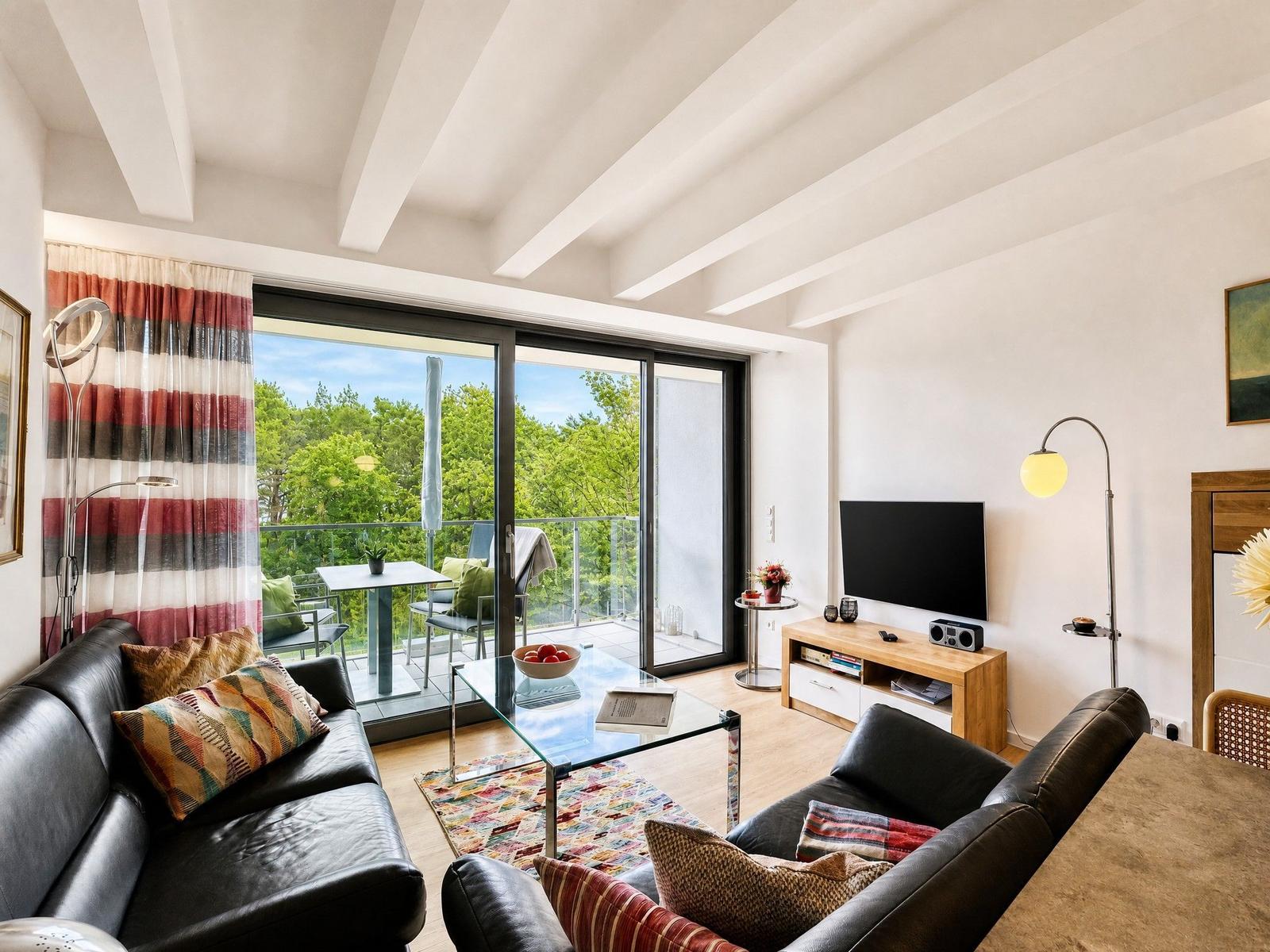 Wohnzimmer mit Sofa, Glas-Tisch und Balkon mit Blick auf Bäume.