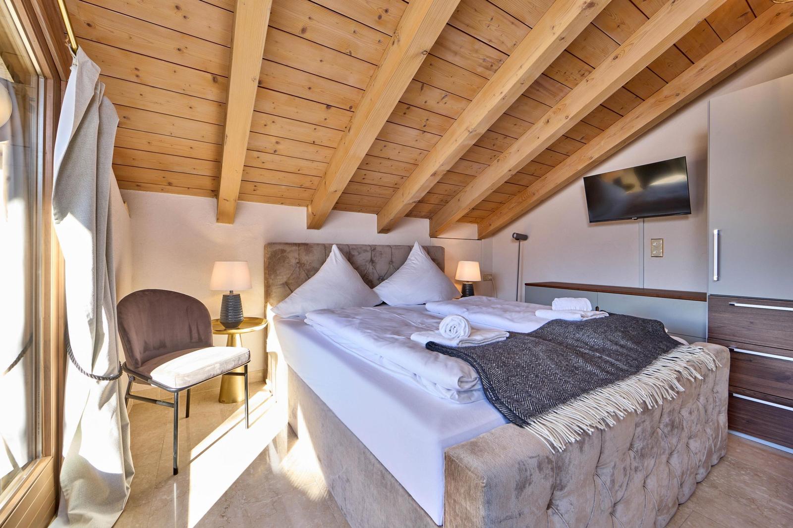 Schlafzimmer mit Bett, TV und Holzdecke. Fenster, Stuhl und Nachttische sichtbar.