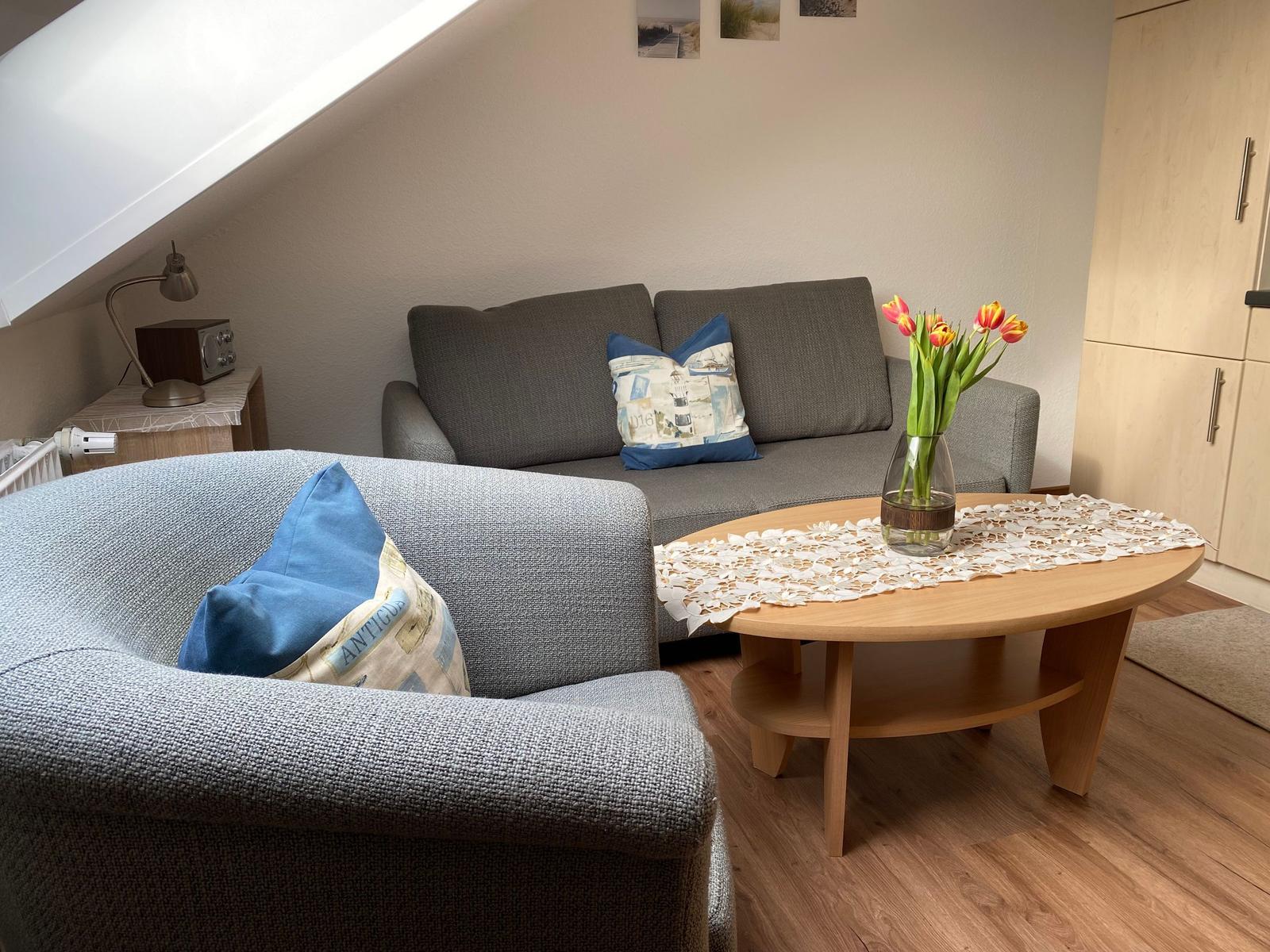 Wohnzimmer mit Sofa, Sessel, Tisch und Blumen. Dachschräge und Holzboden.