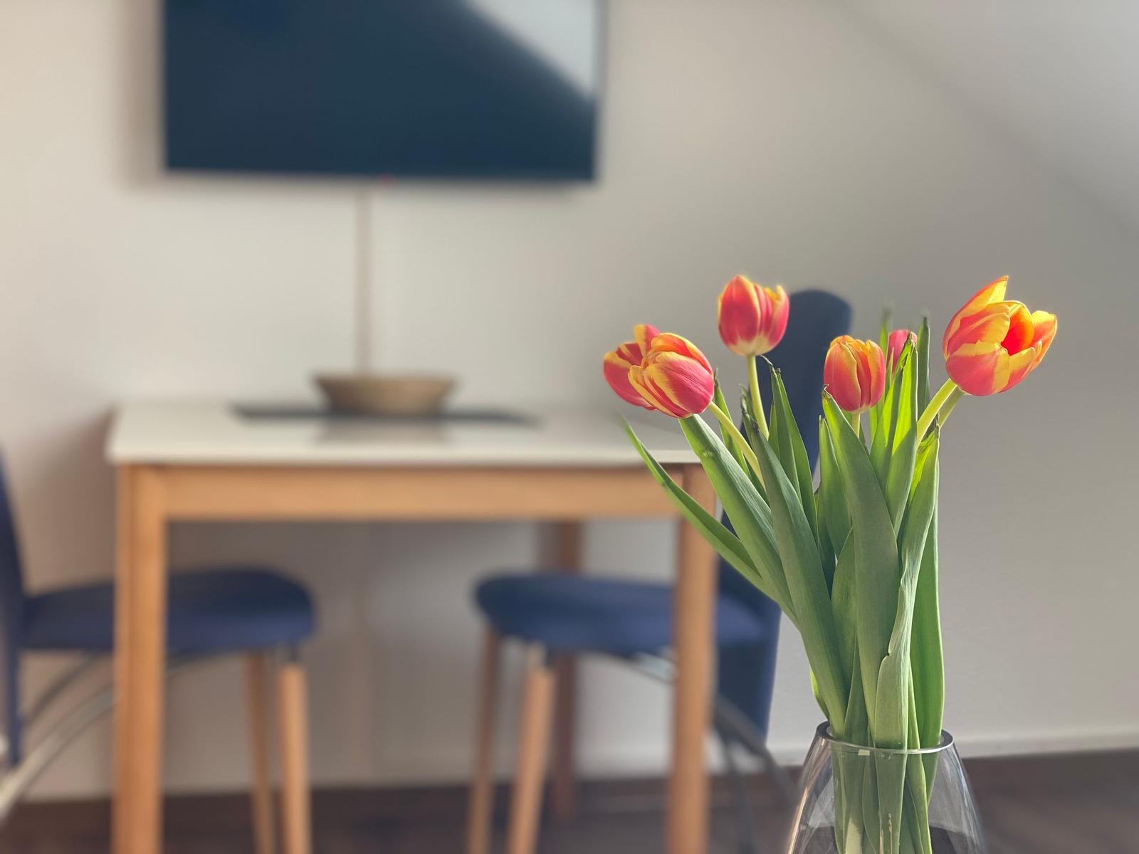 Tulpen in einem Glas stehen vor einem Tisch mit Stühlen und Fernseher an der Wand.