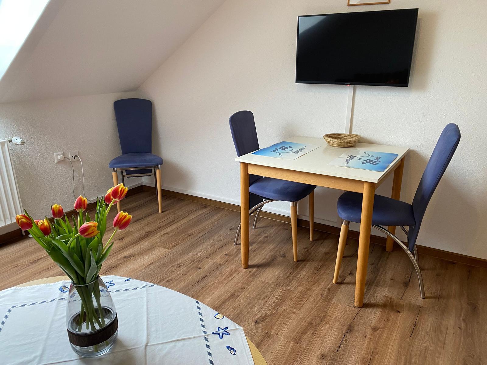 Dreisitzer-Tisch mit blauen Stühlen, Fernseher an der Wand, Tulpen im Vordergrund.