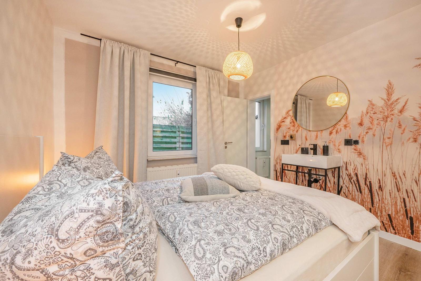 Bedroom with bed, window, mirror, and wall covering with plant motif.