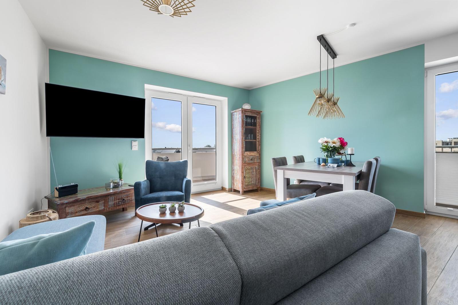 Wohn- und Essbereich mit Fernseher, Sofa und Tisch. Grüne Wände und große Fenster.