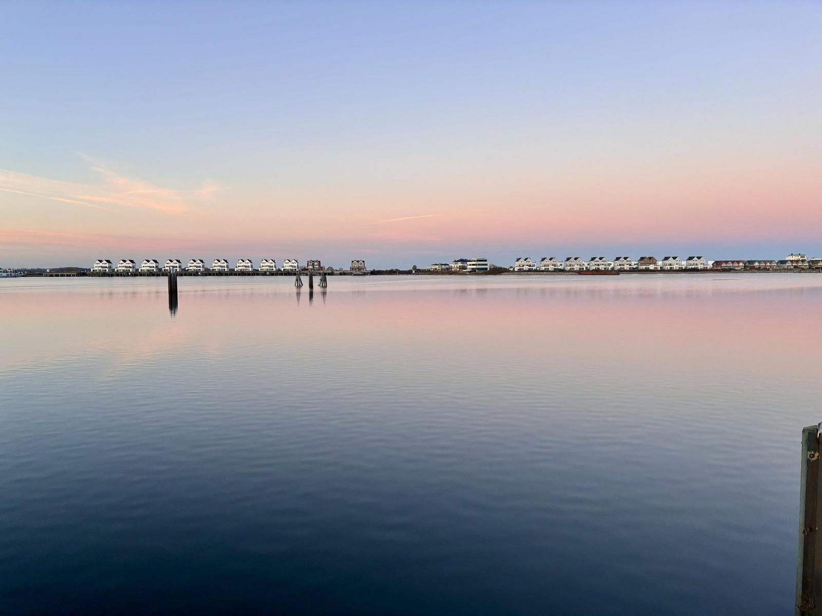 Stille Wasserfläche mit Blick auf Ferienhäuser am Horizont bei Sonnenuntergang.