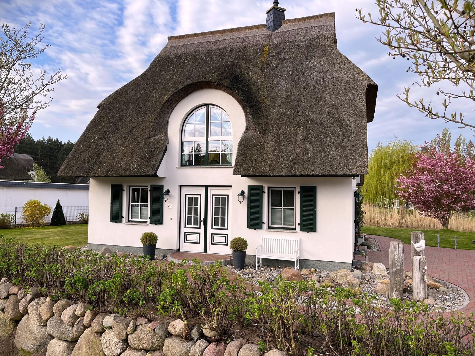 Haus mit Strohdach, weißen Wänden und grünen Fensterläden. Vorgarten mit Steinmauer und Blumen.